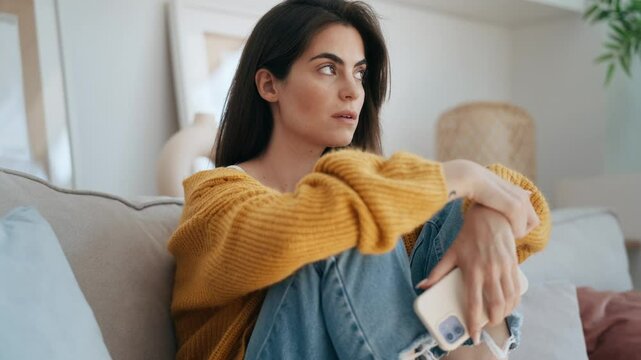 Video of sad young woman overthinking while holding mobile phone sitting on the sofa at home