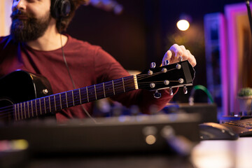 Sound engineer artist tuning his acoustic guitar strings to play and record new tracks, working with electronic mixing and mastering gear in home studio. Young musician creating cool tunes.