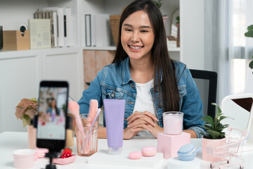 Young beautiful Asian girl reviewing cosmetic and skin care product with new collection's promotion selling on social media online live blurred phone screen surrounded beauty merchandise. Stratagem.