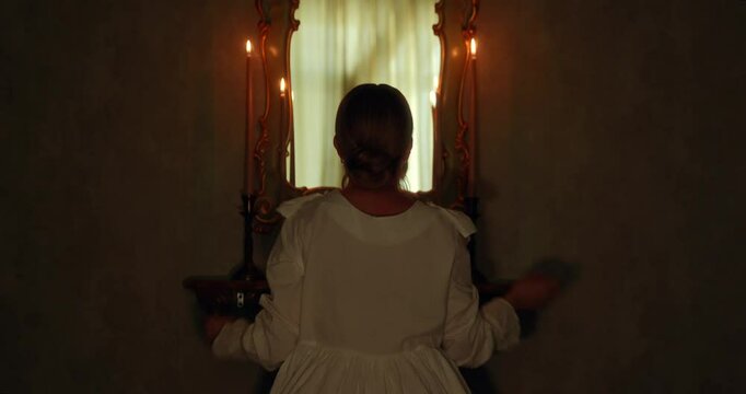 Unrecognizable faces. A view from the back, a young woman in a white nightgown is sitting in a dark room in front of a mirror with candles, raising her hands up and getting ready for bed, 18th century