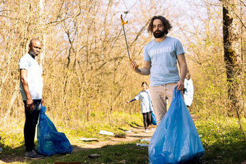 Fototapeta premium Activist using claw tool to grab rubbish and plastic waste from the ground, storing garbage in a trash bag and clean the natural environment. Male environmentalist gathering litter.