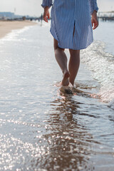 young woman barefoot walks along the sandy beach by the sea at sunset, plunging her feet into the waves and sand, pleasant relaxation on summer vacation