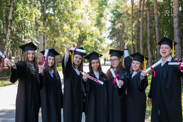 Row of young students in graduation gowns outdoors showing off their diplomas. 