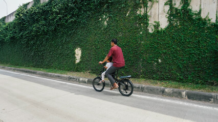 Obraz premium A father and son riding a bicycle on a quiet road, flanked by a tall green ivy wall. A serene urban scene capturing familial joy