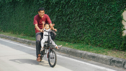 Obraz premium A father and son joyfully riding a bicycle along an ivy-covered wall in a quiet urban area. The warmth of family in the city