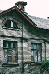 Exterior view of an old one-story white brick house with an attic and a ventilation outlet on the roof. A 20th century stone building with windows that are covered with various objects from the inside