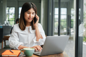 A young professional woman multitasking by working on her laptop and talking on the phone in a modern office setting.