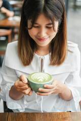 A young woman holding a cup of matcha latte, smiling and enjoying her drink in a cozy cafe setting.