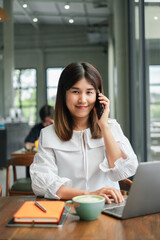 Obraz premium A young woman in a white blouse working on a laptop and talking on the phone in a modern cafe, with a coffee cup and notebook on the table.