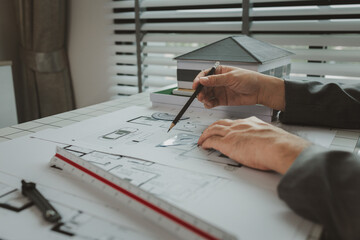 An architect is working in the office, there are blueprint and house model on the desk, engineer is checking overall of work that going to be present one last time, using tool for making work easier