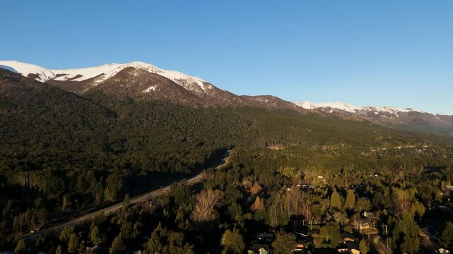 This breathtaking aerial photograph captures the scenic beauty of Cerro Bayo, a mountain located in Villa La Angostura, Argentina. The image showcases a vibrant forested landscape with snow-capped 