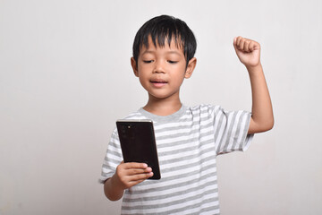 A happy and excited Asian boy with raised hands looking at smartphone isolated on white background. little boy looking at phone. Technology concept, copy space