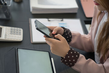 An Asian businesswoman checks her smartphone while working at her desk, surrounded by a laptop, documents, and stationery in a modern office
