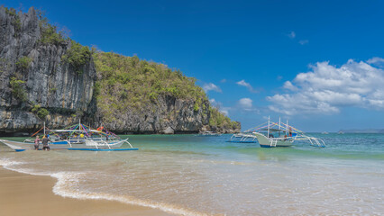 Traditional Filipino bangka boats are anchored. The waves of the turquoise ocean spread over the sandy beach. Silhouettes of people. Sheer karst cliffs with green vegetation against the blue sky