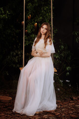A caucasian woman in an elegant white lace dress sits on a wooden swing amidst dark foliage. 
