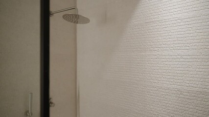Modern shower with glass door partially open. Interior with white textured tiles and large showerhead mounted on ceiling. Soft and ambient light creating calm atmosphere