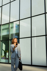 Business woman strolls confidently outdoors in smart attire, checking her messages against the backdrop of a modern glass building on a sunny day