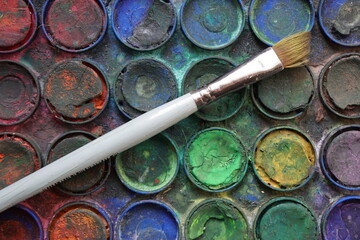 Used paintbrush placed on a set of dirty colorful watercolors in a palette
