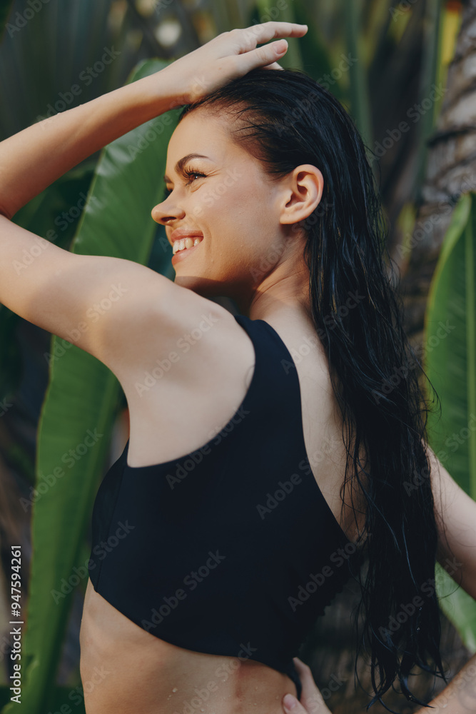 Wall mural beautiful woman in black bikini top posing in front of palm tree with hand on head on tropical beach