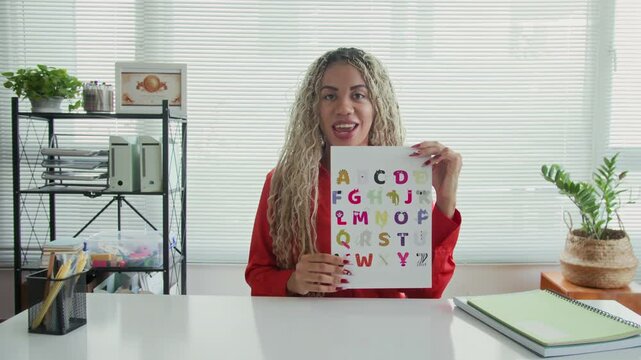 Medium POV of young female Latin speech therapist talking on camera about correct pronunciation of letters when sitting at desk in room