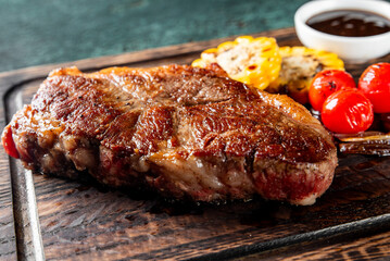 Grilled steak on a wooden cutting board, garnished with corn, cherry tomatoes, garlic cloves, and a bowl of sauce. Perfect for a barbecue or culinary presentation.