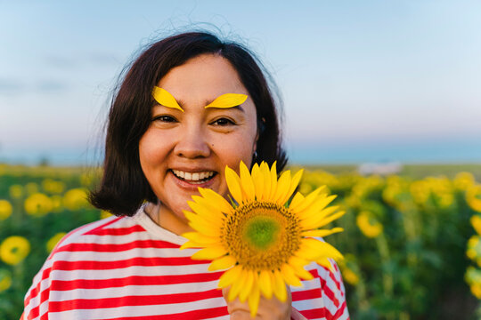 Happy woman with sunflower wearing petals on eyebrows in field