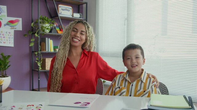 Medium portrait of female Hispanic logopedist and Asian schoolboy looking at camera when sitting at desk in classroom