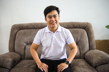 Cheerful Asian Man Sitting On Sofa While Smiling At Camera