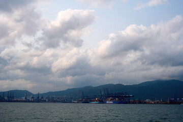 A cloudy sky looms over Hong Kong's busy port with cranes and mountains, showcasing a blend of...
