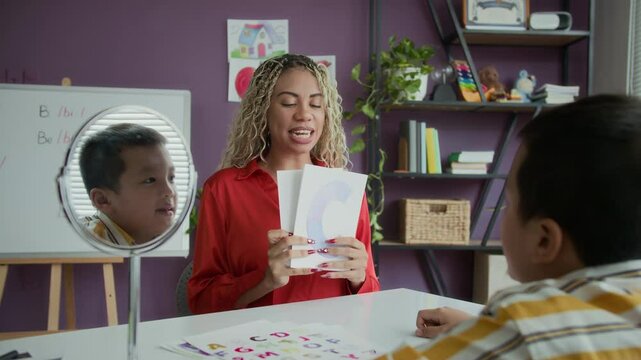 Latin female tutor practicing pronunciation of letters with teenage Asian boy looking at himself in mirror at classroom