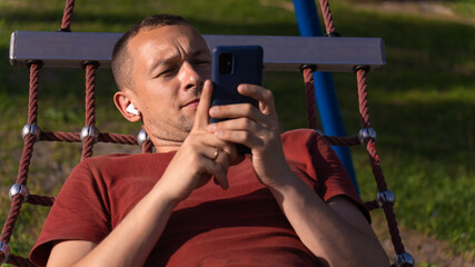 Obraz premium Portrait of a man looking at a mobile phone lying in a hammock and listening to music through white wireless headphones in the park.
