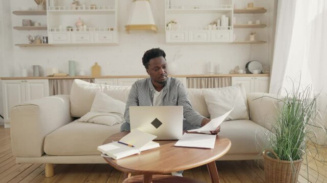 African American man working from home remotely, sitting on sofa, using laptop. Portrait of young adult freelancer man at home, workplace and home office of employee, black male person with computer