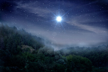 Bright star indicates the Nativity of Jesus Christ in the starry sky and fog is rising over the night forest