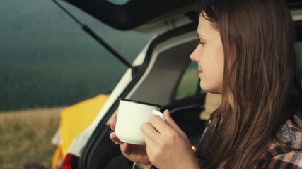 Happy traveller woman sitting in open trunk of car and drink hot coffee on background mountain and yellow tent at summer or autumn morning. Hiking, adventure active vacations, camping vibes concept