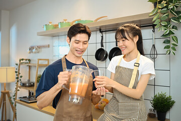 Beautiful young couple wearing apron enjoying healthy fruit smoothie in domestic kitchen
