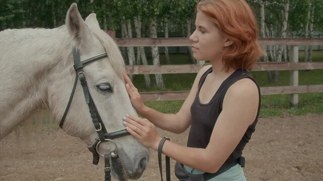 Young red-haired Caucasian woman standing in outdoor practice arena and stroking muzzle of beautiful white horse with tenderness