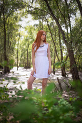 Redhead girl in the white dress posing in the forest thicket.