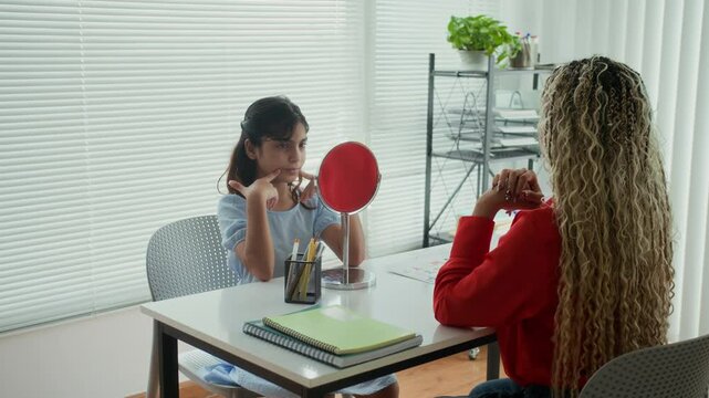 Female tutor guiding Indian schoolgirl pronouncing sounds in front of mirror at speech therapy class