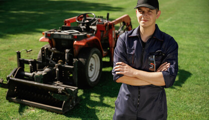 Standing with arms crossed. Man is with utility tractor with grass cutter and aerator equipment on...
