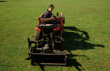 Obraz premium Driving, looks behind, shows thumb up. Man is with utility tractor with grass cutter and aerator equipment on the field