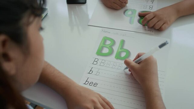 Handheld shot of two children writing letter B on worksheet during logopedic session at school