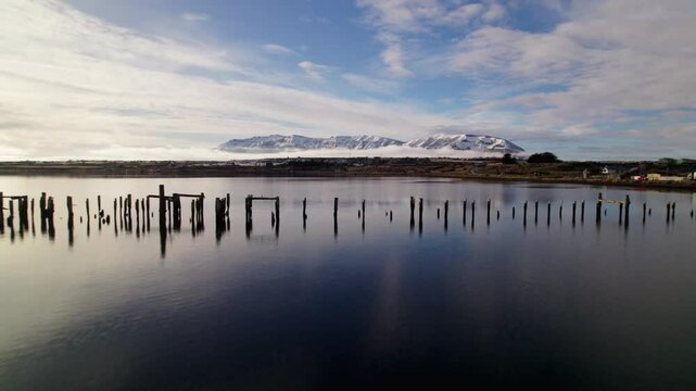 Wide aerial pushback above a blue reflective lake as birds fly past wooden posts, showcasing the snowy misty mountains of Patagonia, Chile.