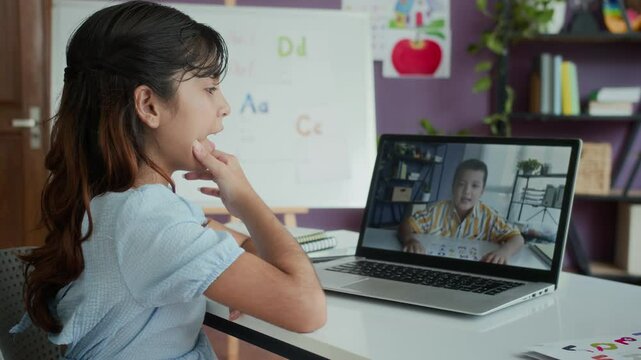 Side view of Indian schoolgirl pronouncing letters of alphabet together with her friend during online class via laptop at table