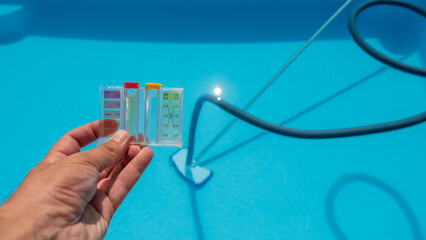 Hand holding a pool water testing kit above a clear blue pool with cleaning equipment in the...