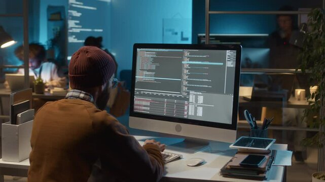 Timelapse back view of unrecognizable male software engineer wearing beanie sitting at computer writing new feature for web application. Workflow of team of IT specialists in background