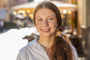 Portrait of happy redhead teenager girl smiling friendly, glad expression looking at camera dreaming, resting, relaxation feel satisfied good news outdoors. Young woman in urban city sunshine street