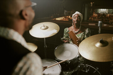 Over shoulder shot of young Black female singer standing in front of drummer singing song and playing guiro