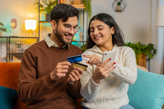 Happy Hispanic couple together doing online shopping by entering credit card number on smartphone while sitting on sofa at home. Smiling family satisfied with a good promotion on goods in apartment.