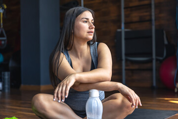 A pregnant woman in the gym sits on a mat and drinks water.