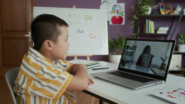 Side view of Asian teenage boy studying online with female logopedist on wireless laptop in classroom